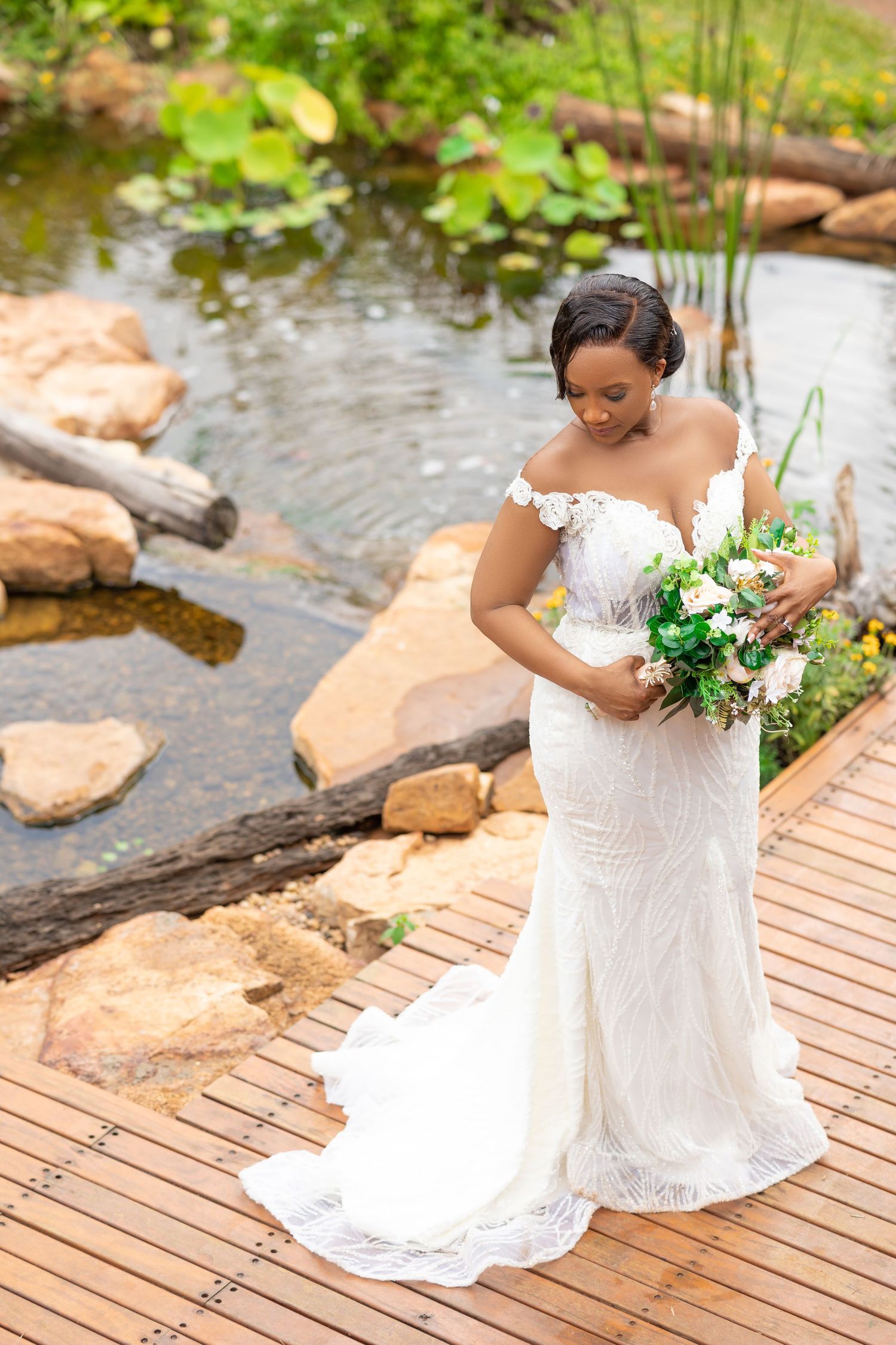 Beautiful bride in a white lace wedding dress poses with her bouquet on a wooden deck by a scenic pond.