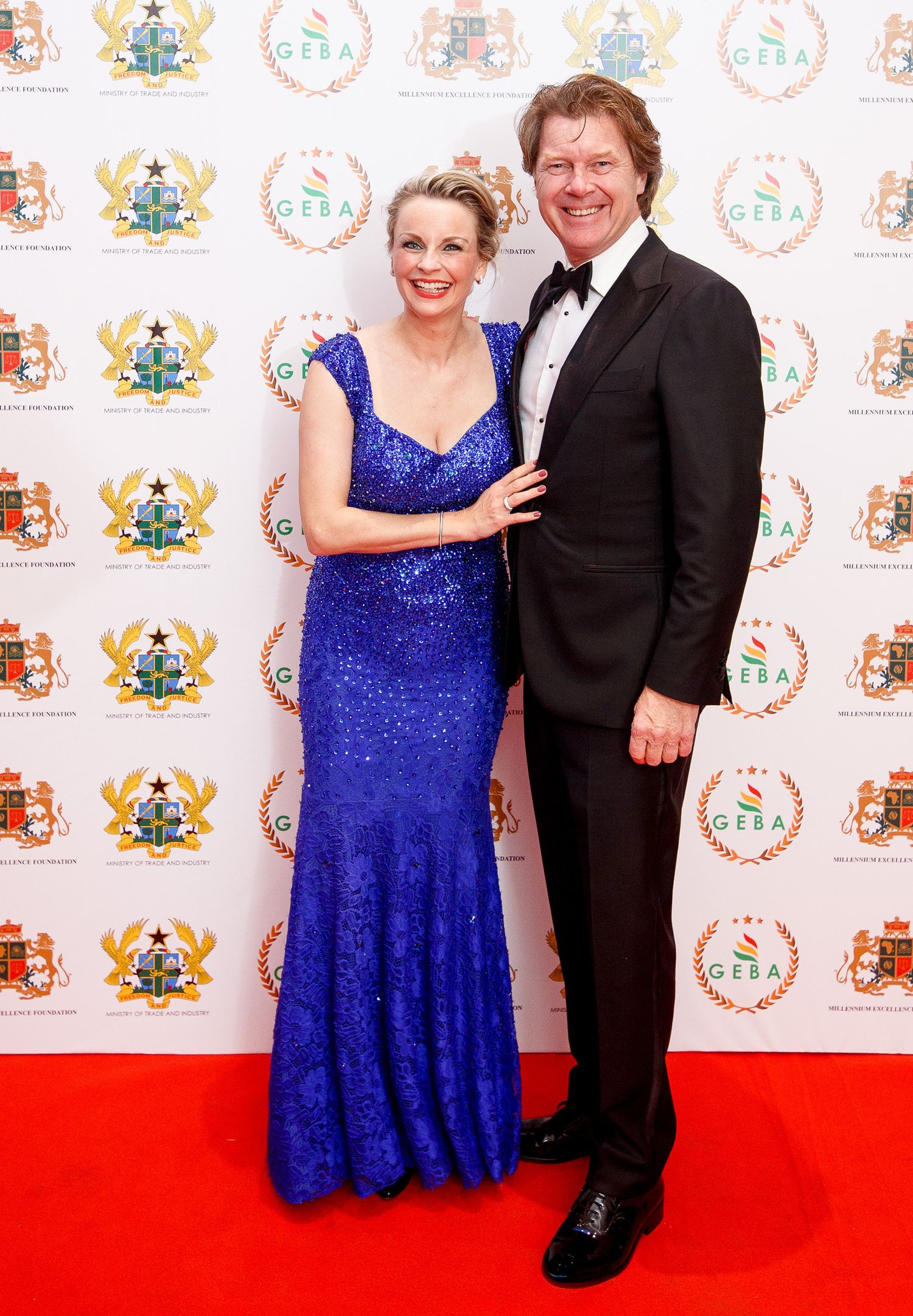 A couple poses together on a red carpet, with the woman wearing a sparkling royal blue gown and the man in a black tuxedo.