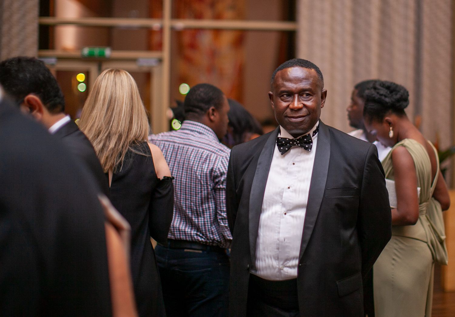 Guests in formal attire mingle at an elegant evening event with warm lighting and decorative columns in the background.
