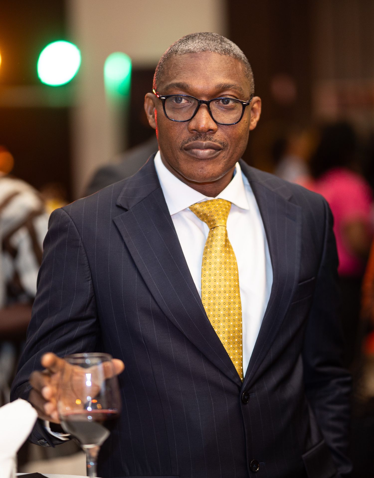 A person in a dark business suit and yellow tie smiles while holding a drink at an evening event.