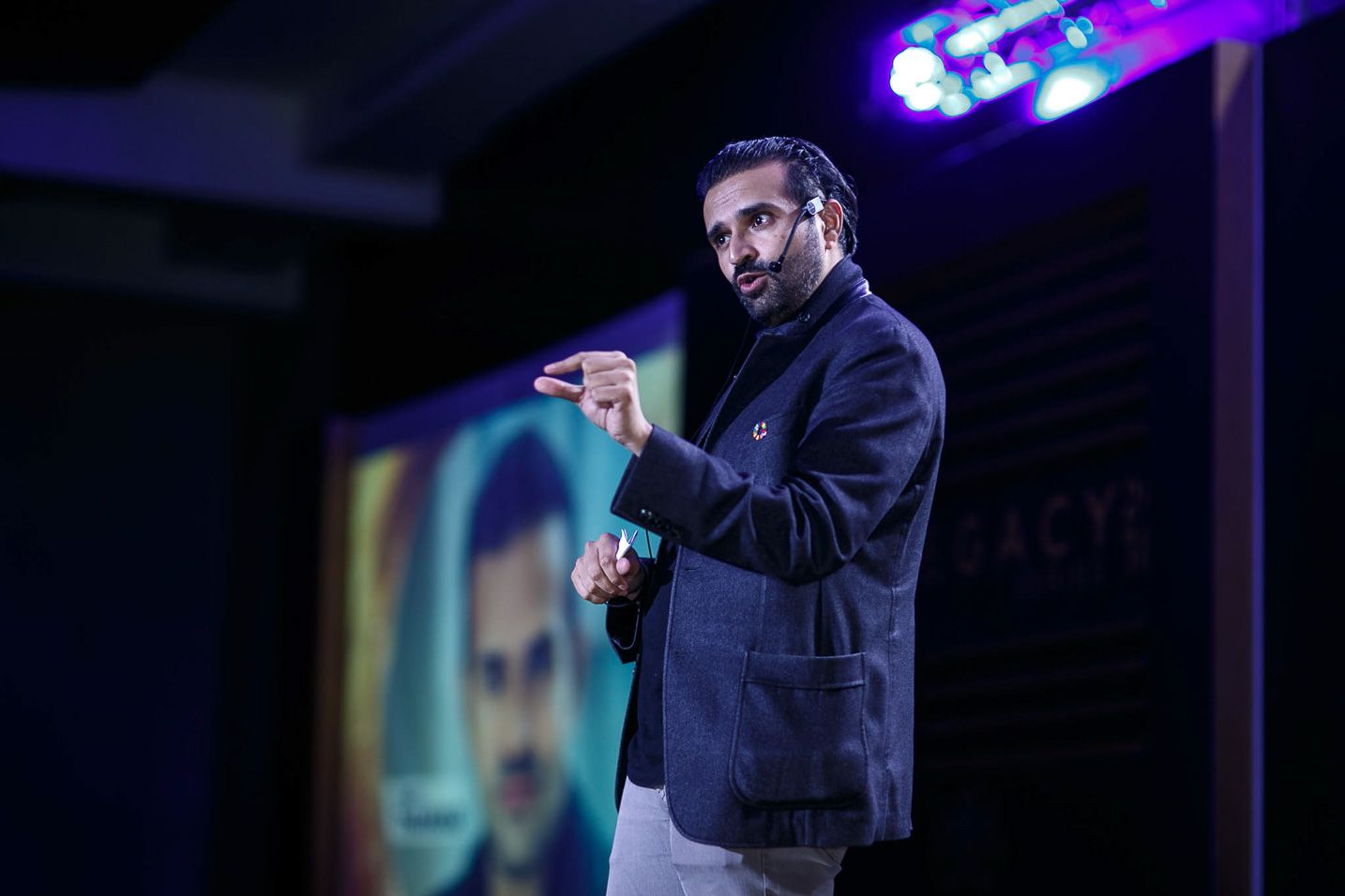 A performer in a dark suit gestures expressively while speaking on stage with blue and purple lighting effects.