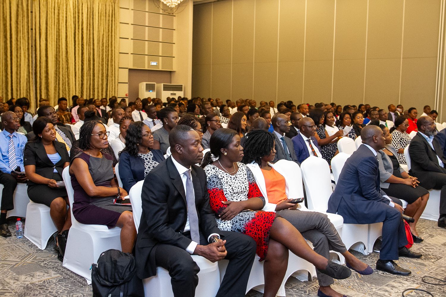 A large audience sits attentively in rows of chairs at a professional conference or business event.