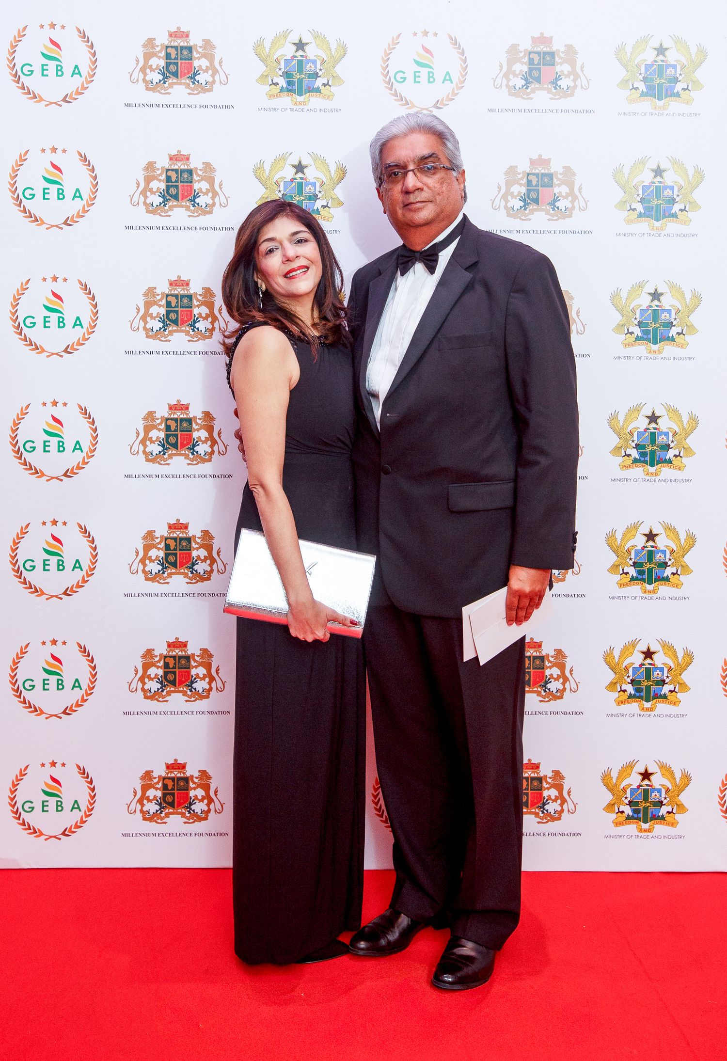Two people in formal evening attire pose together on a red carpet with repeating logo backdrop at an event.