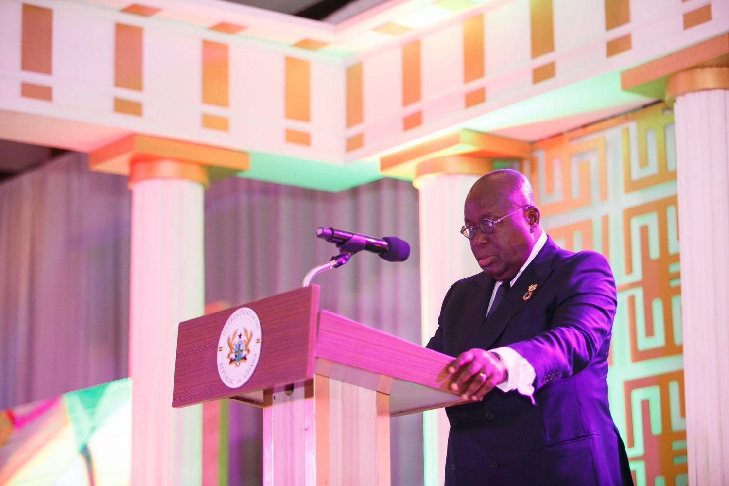 A speaker stands at a podium with colorful lighting and architectural details in the background.