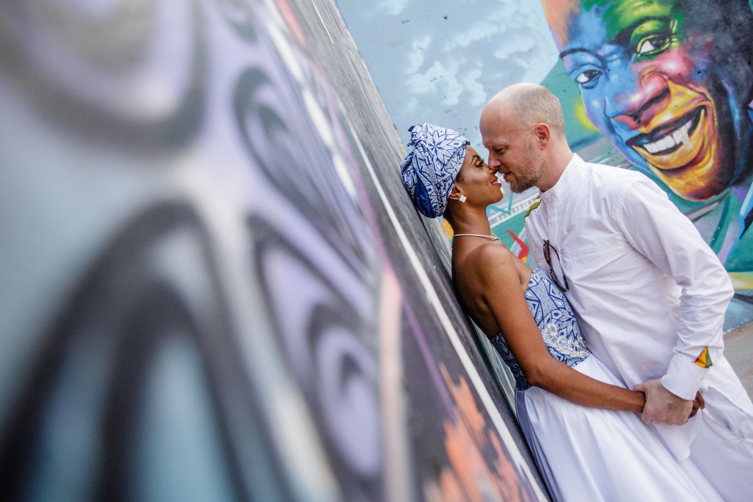 A couple shares a romantic moment against a vibrant graffiti wall featuring a colorful painted face.
