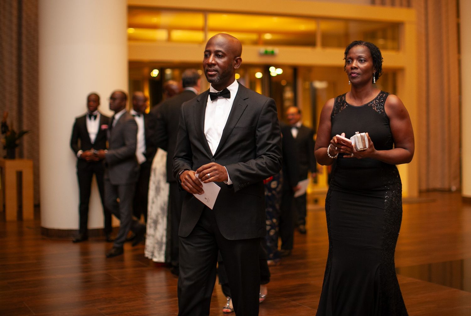 Guests in formal black tie attire mingle and socialize in an elegant hotel hallway during an evening event.