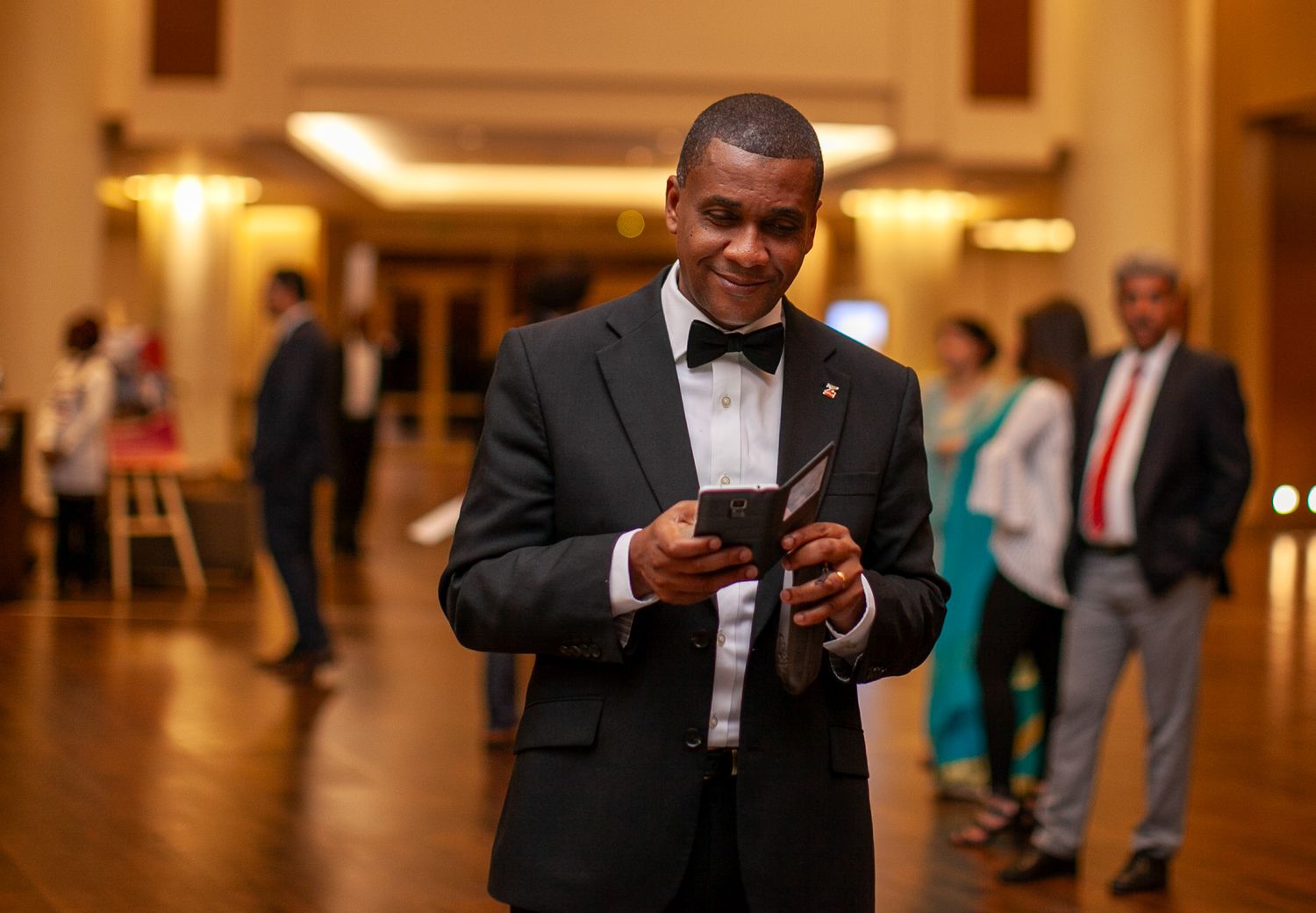 A speaker in formal attire holds a microphone while presenting in a warmly lit hotel conference space.