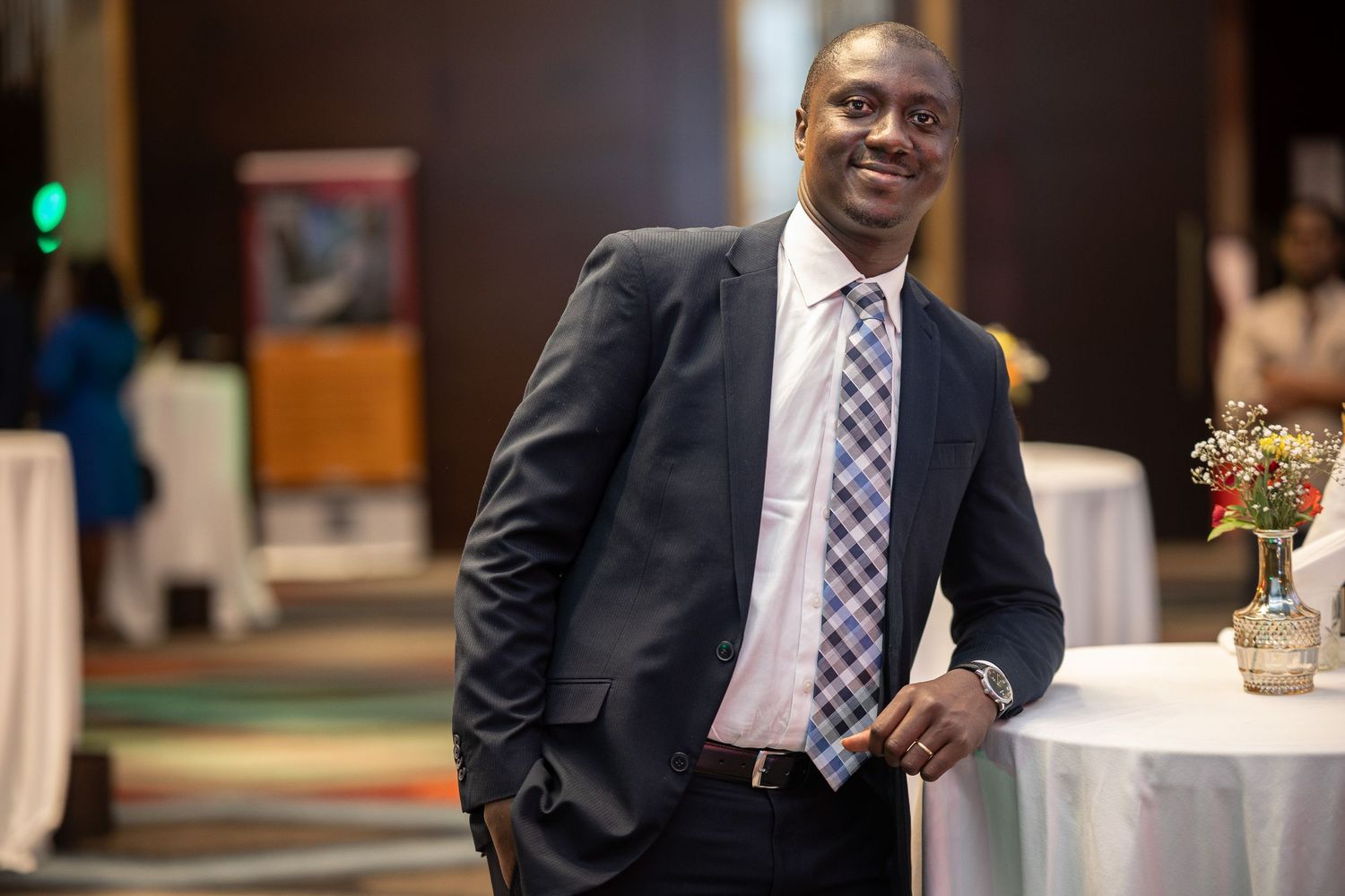 Professional in a navy suit and checkered tie stands at an elegant event with ambient lighting.