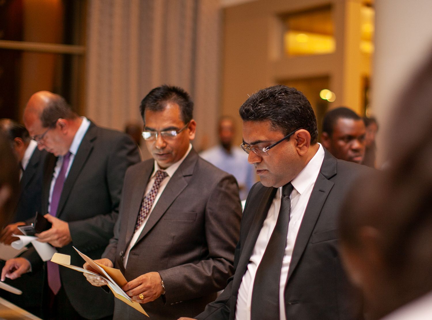 Business professionals in dark suits gather at a networking event, checking their mobile phones in a hotel lobby.