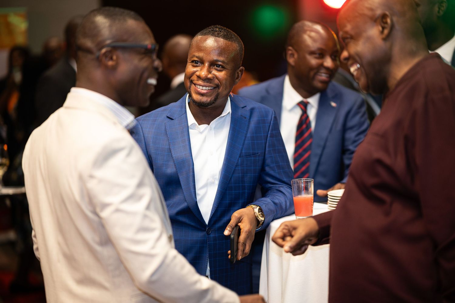 A group of men in suits and formal attire laugh and smile together at a social gathering.