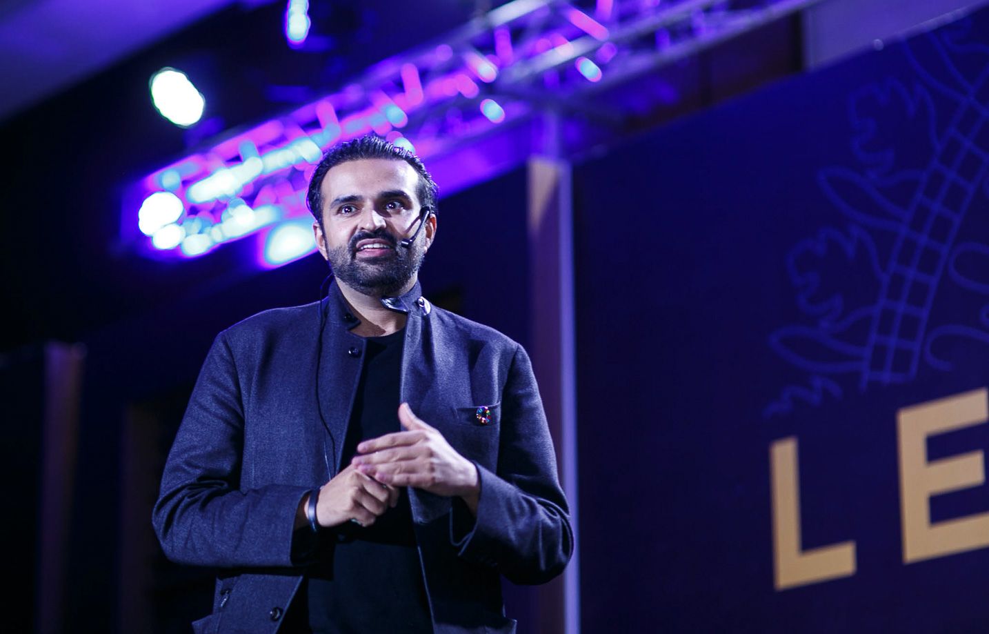 Speaker on stage with blue lighting and technical equipment visible in the background.