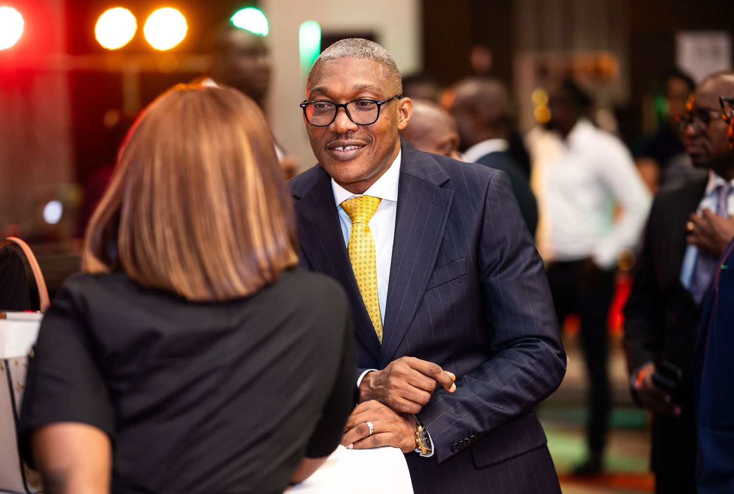 Professional in glasses and yellow tie smiles warmly during networking event with blurred attendees in background.