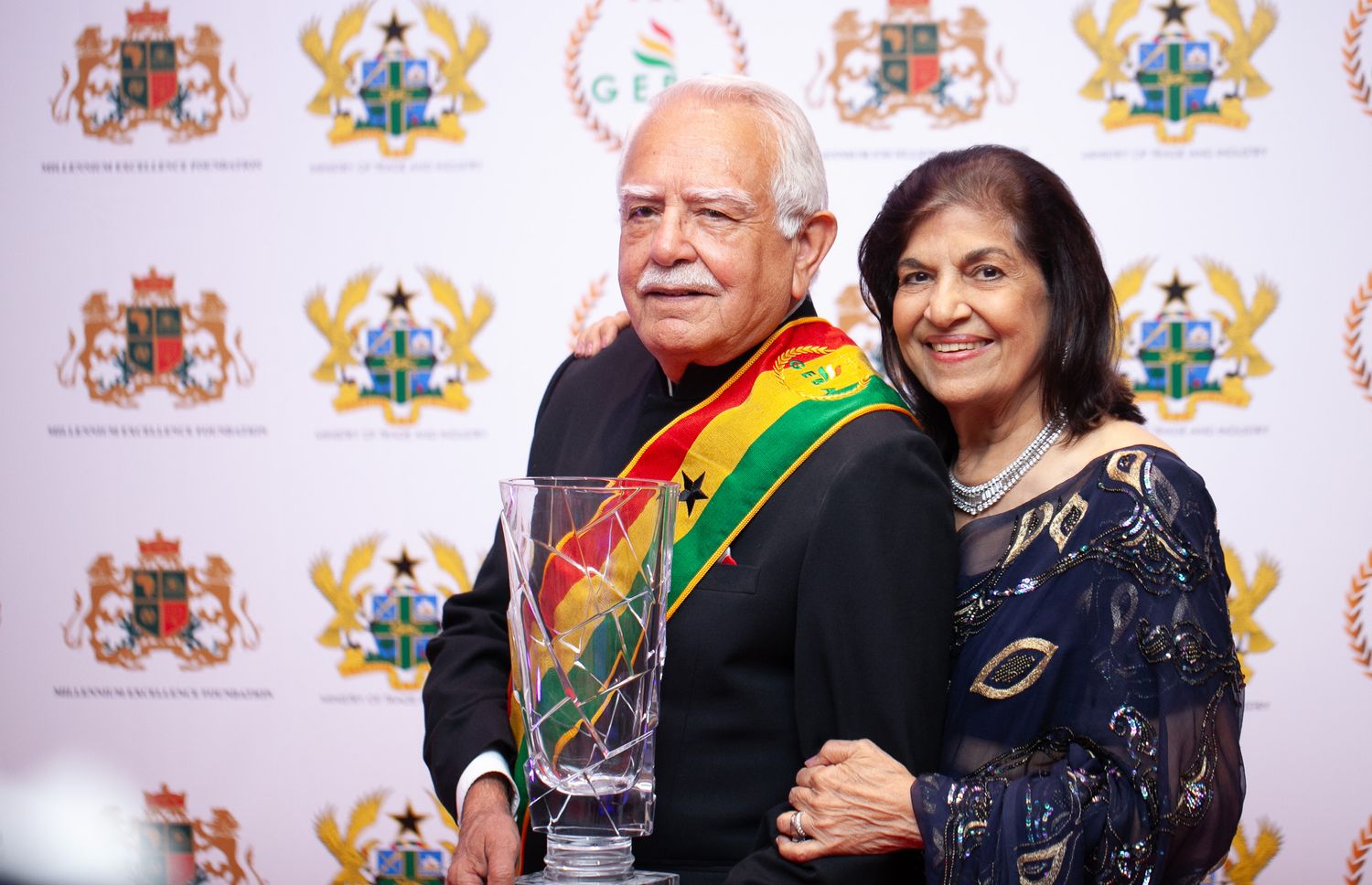 Two people smile while posing together at an event with official emblems and flags visible in the background.