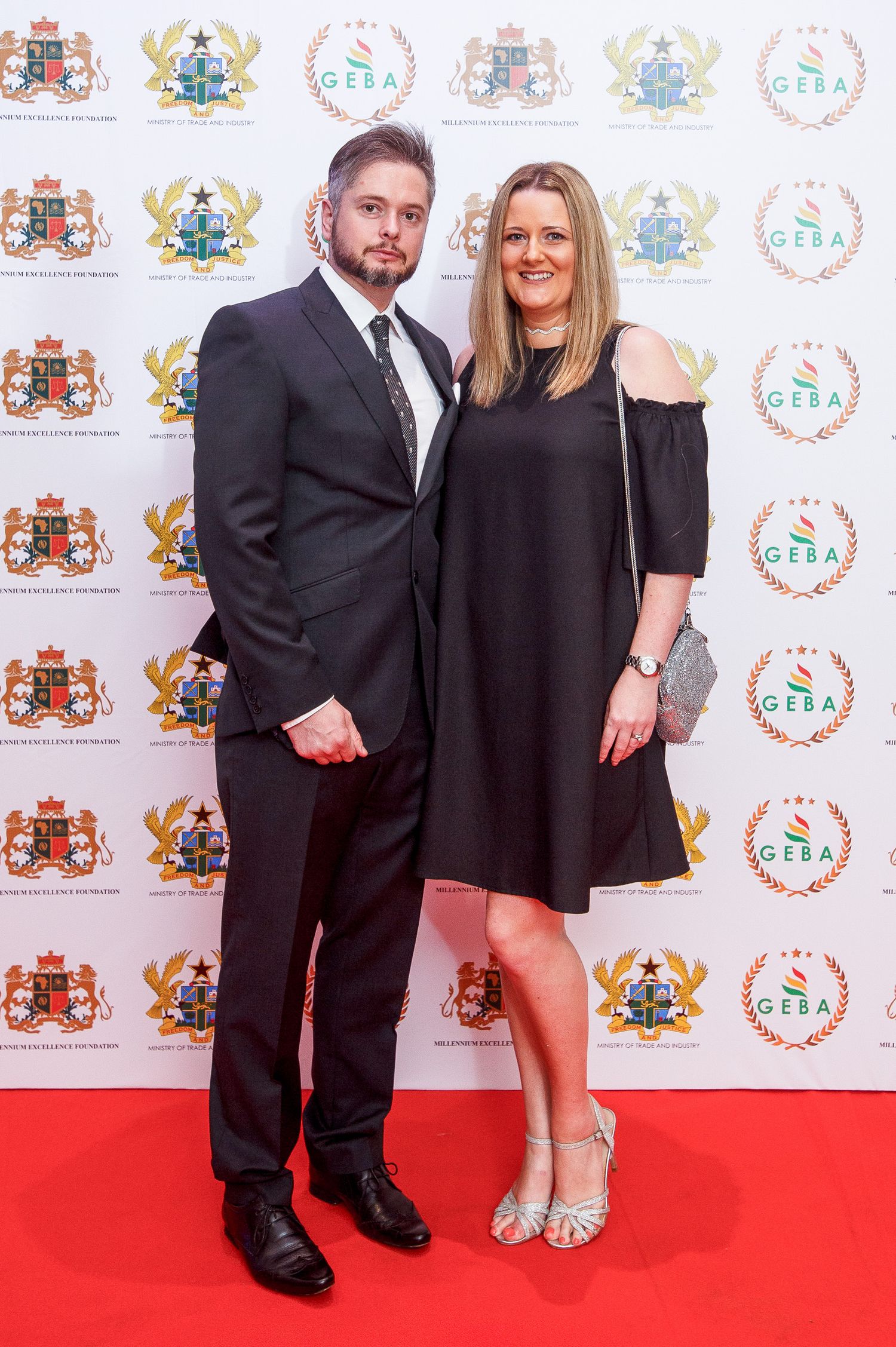 A couple poses together on a red carpet at a formal event with logo-decorated backdrop.