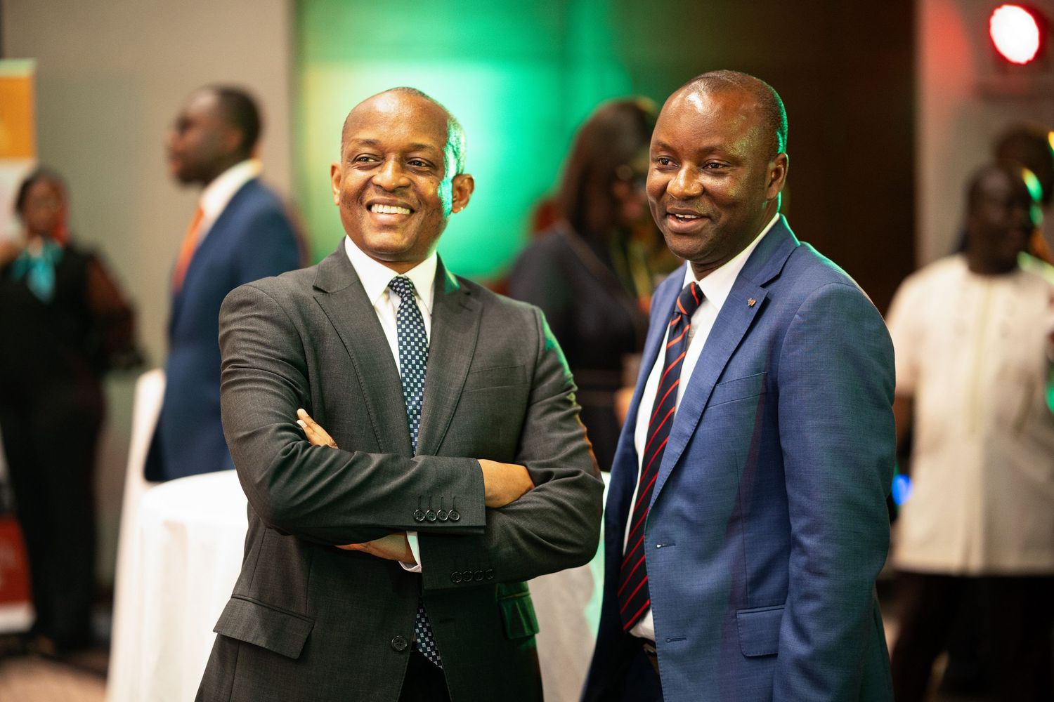Two businessmen in suits share a friendly moment at a professional event with green lighting in the background.
