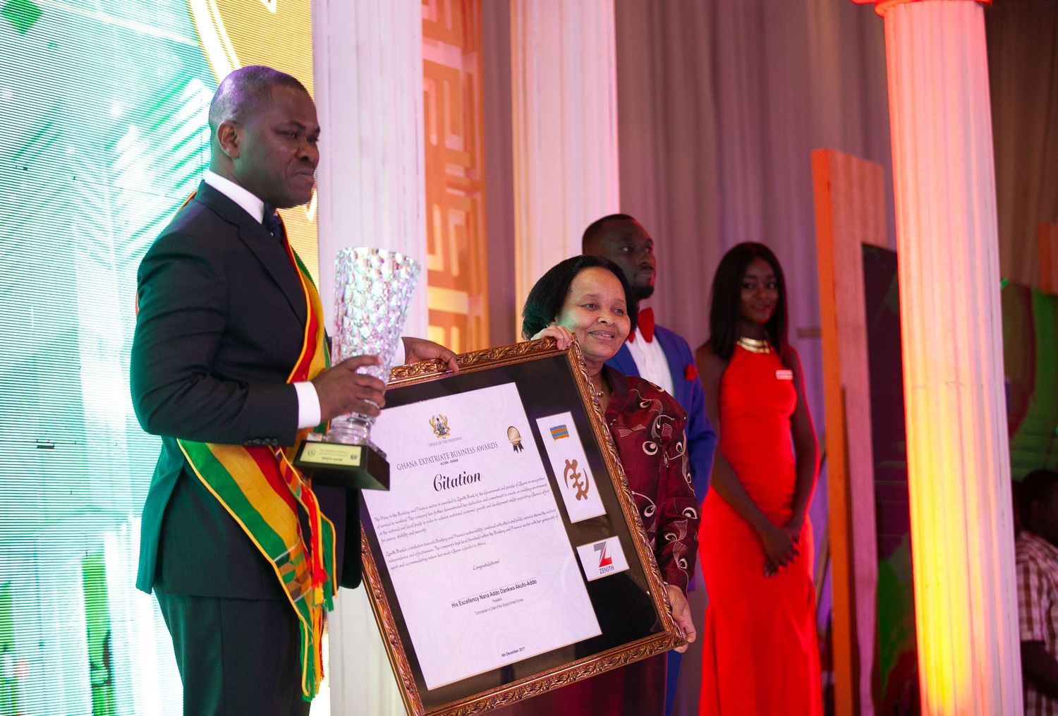 Graduates in red and black academic regalia stand in line during a formal ceremony with green lighting effects.