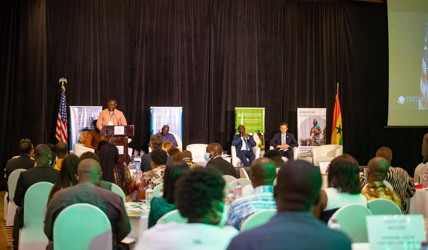 Conference panel discussion with speakers seated at a long table on stage with audience members in white chairs.