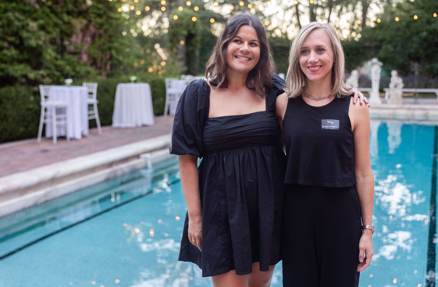 Two people wearing black outfits pose together by a swimming pool on a sunny day.