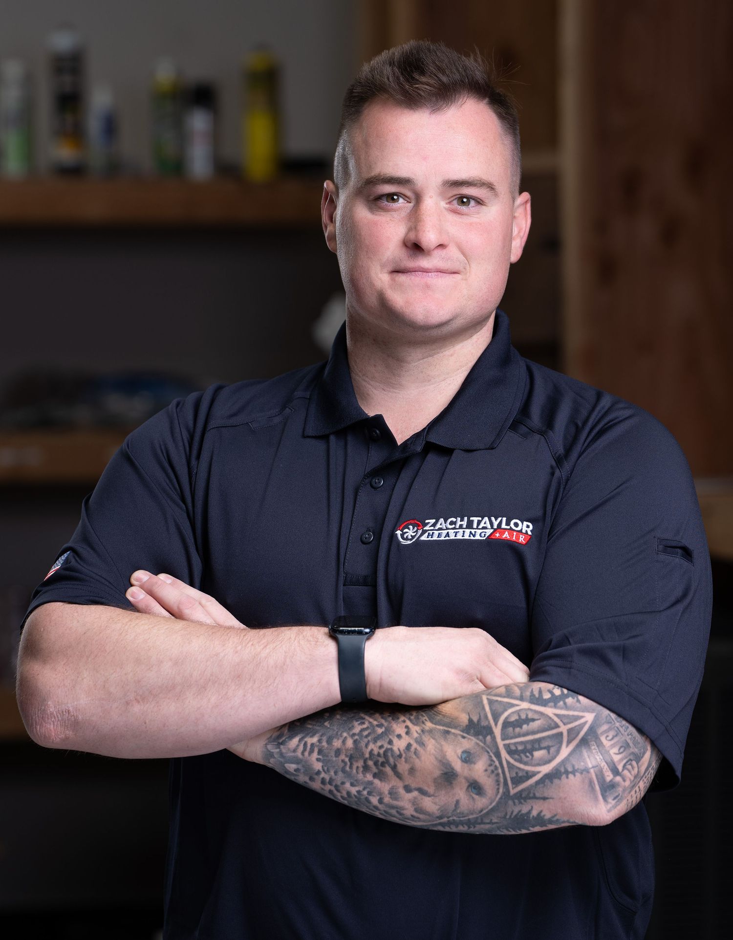 Professional portrait of someone in a black polo shirt showing tattooed arms while posing against a blurred background.
