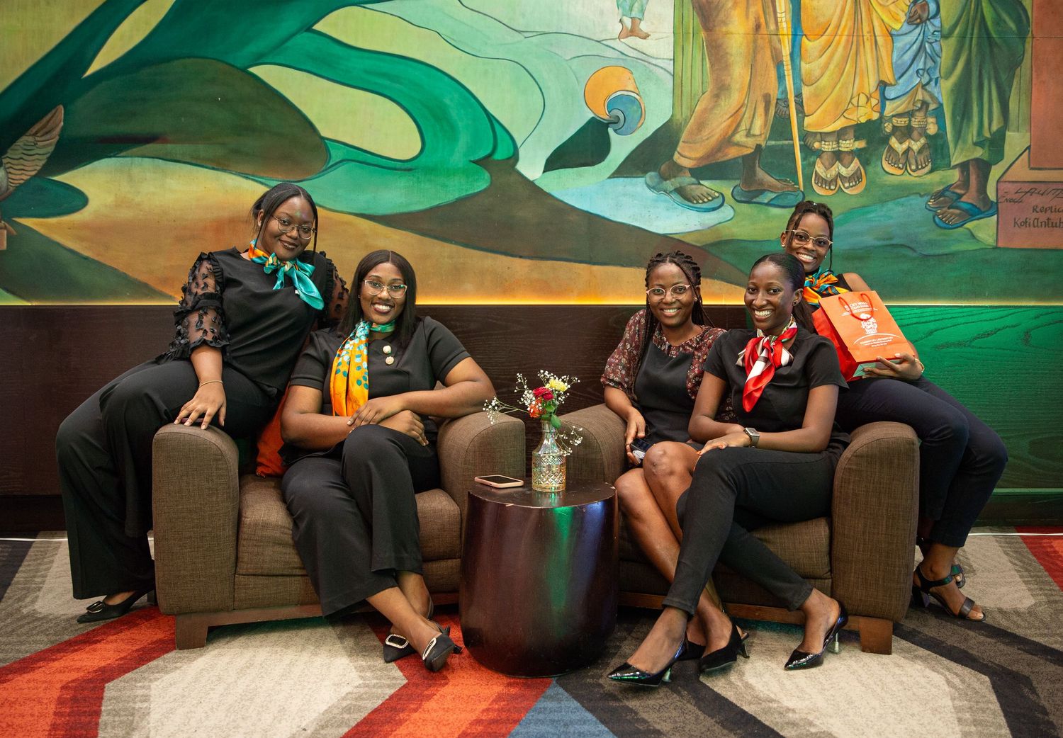 A group sits together on a brown couch, laughing and socializing in front of a vibrant green mural.