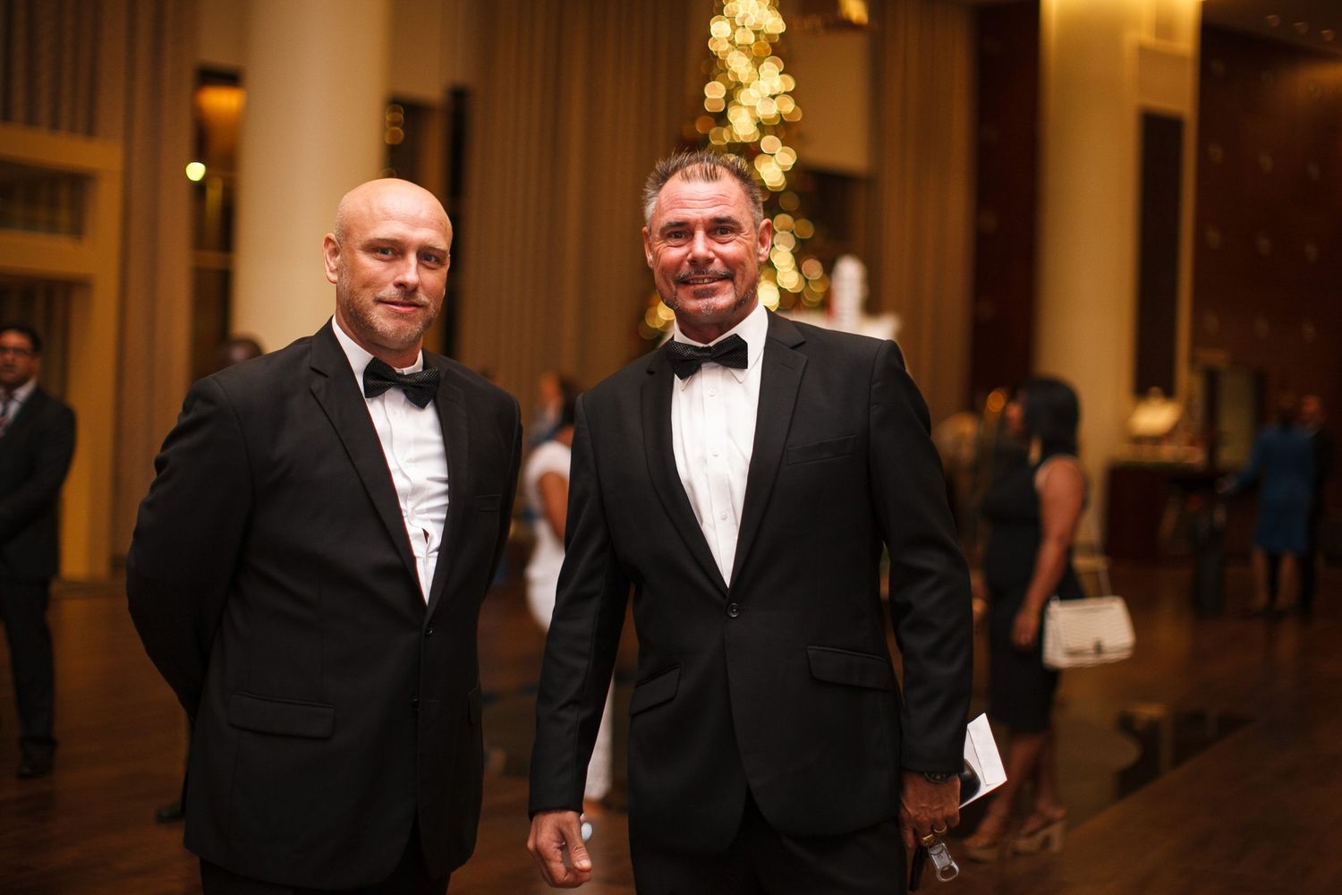 Two people in formal black tuxedos and white shirts stand smiling in an elegant hallway with warm lighting.