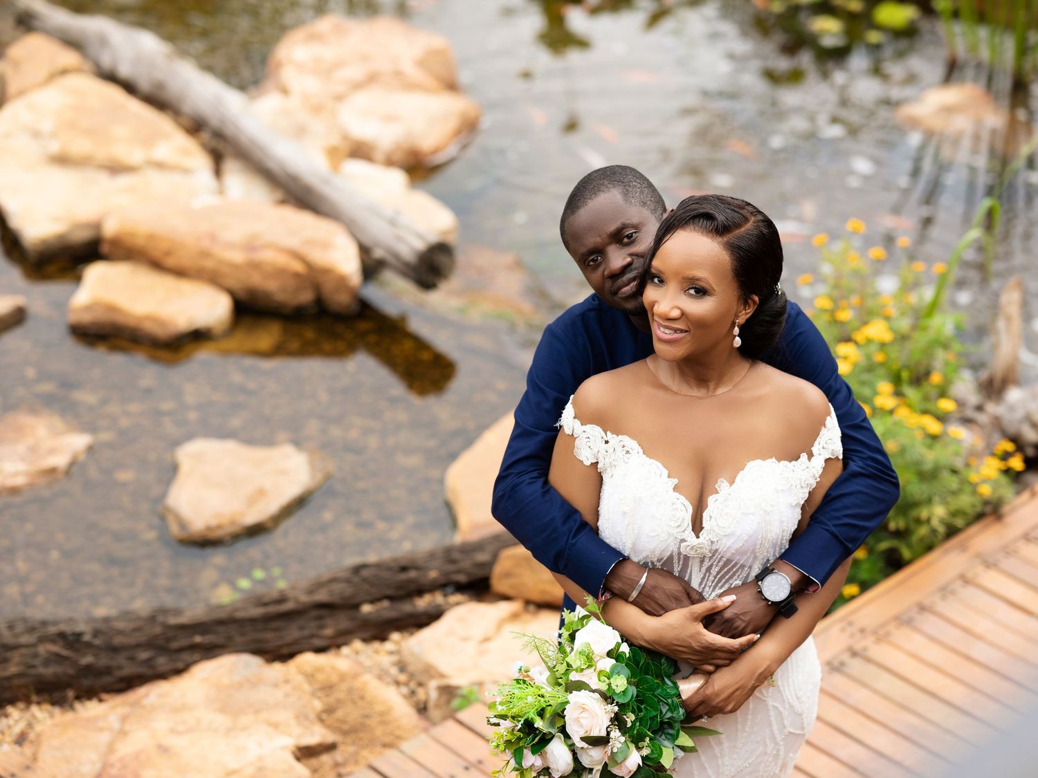 A couple shares an intimate moment during their outdoor wedding photoshoot near a serene pond with rocks.