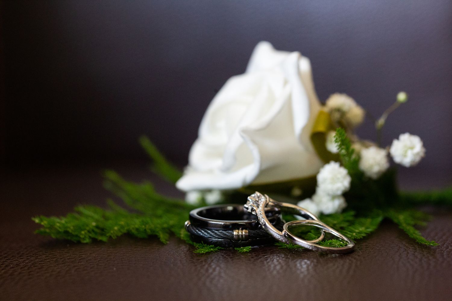 Wedding rings and delicate white flowers arranged on dark surface with soft lighting and greenery accents.