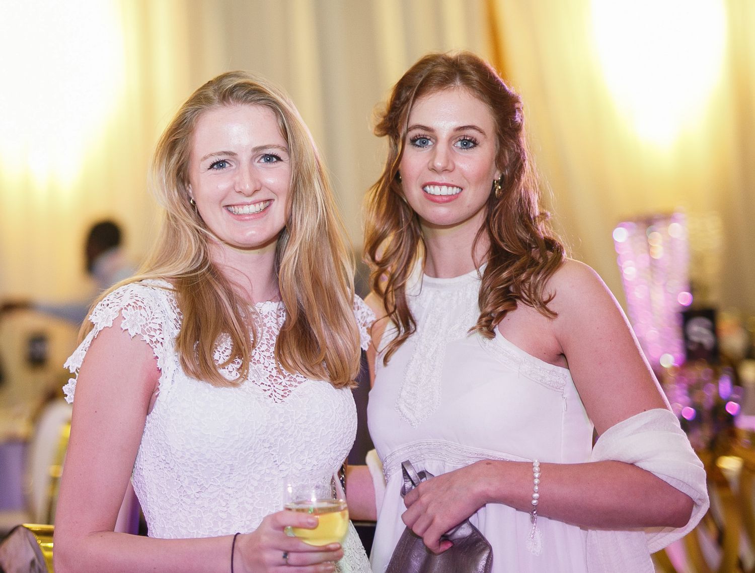 Two women in white dresses smile while attending a formal event with a warm glowing background.