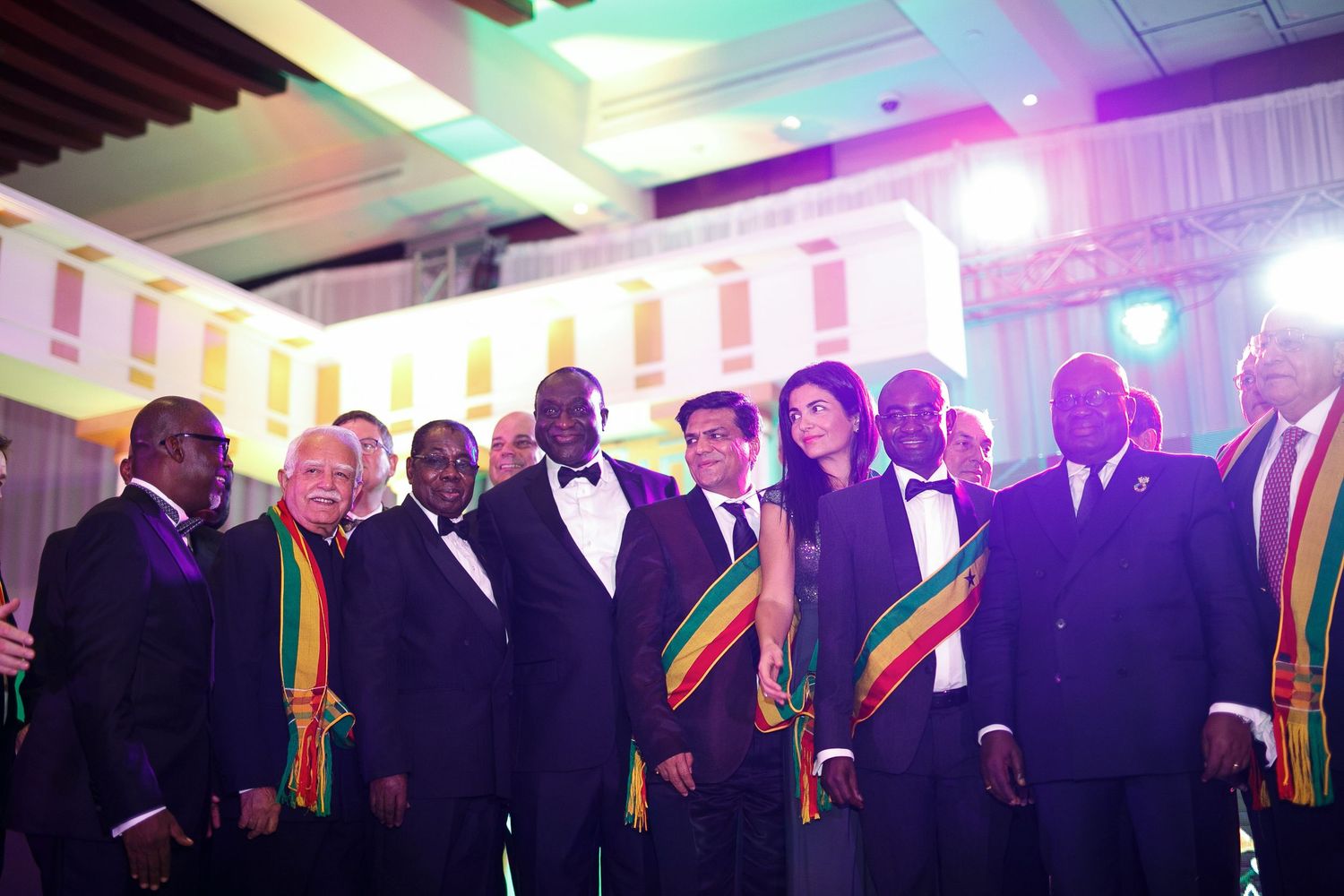Group of people wearing rainbow-striped sashes stand together in a formal indoor setting with colorful lighting.