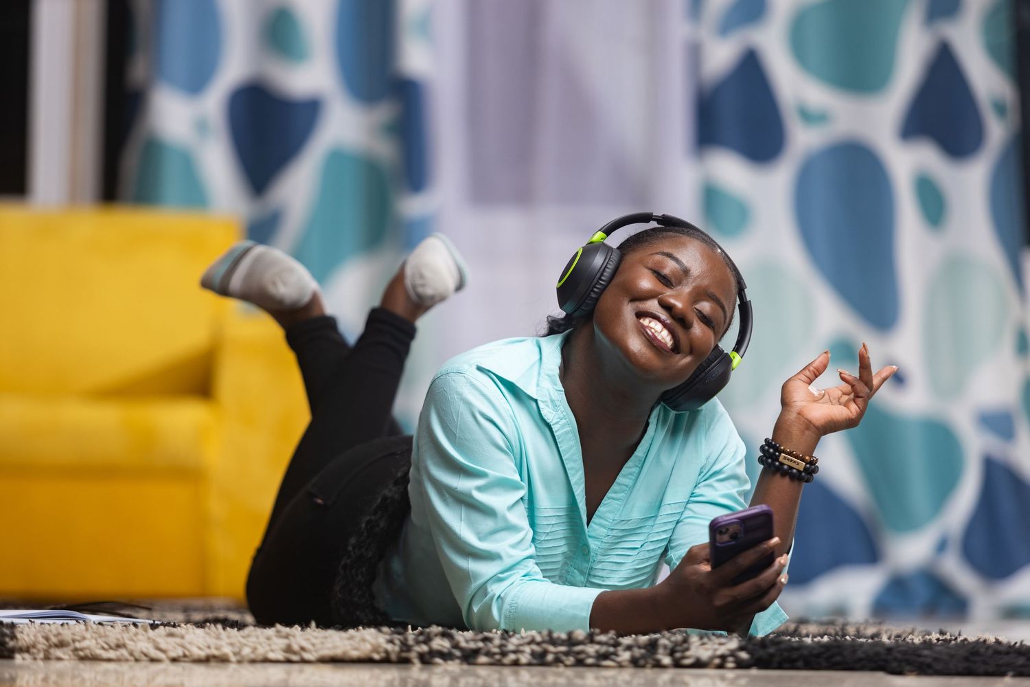 Someone lying on floor listening to music with headphones while relaxing by yellow couch with patterned curtains.