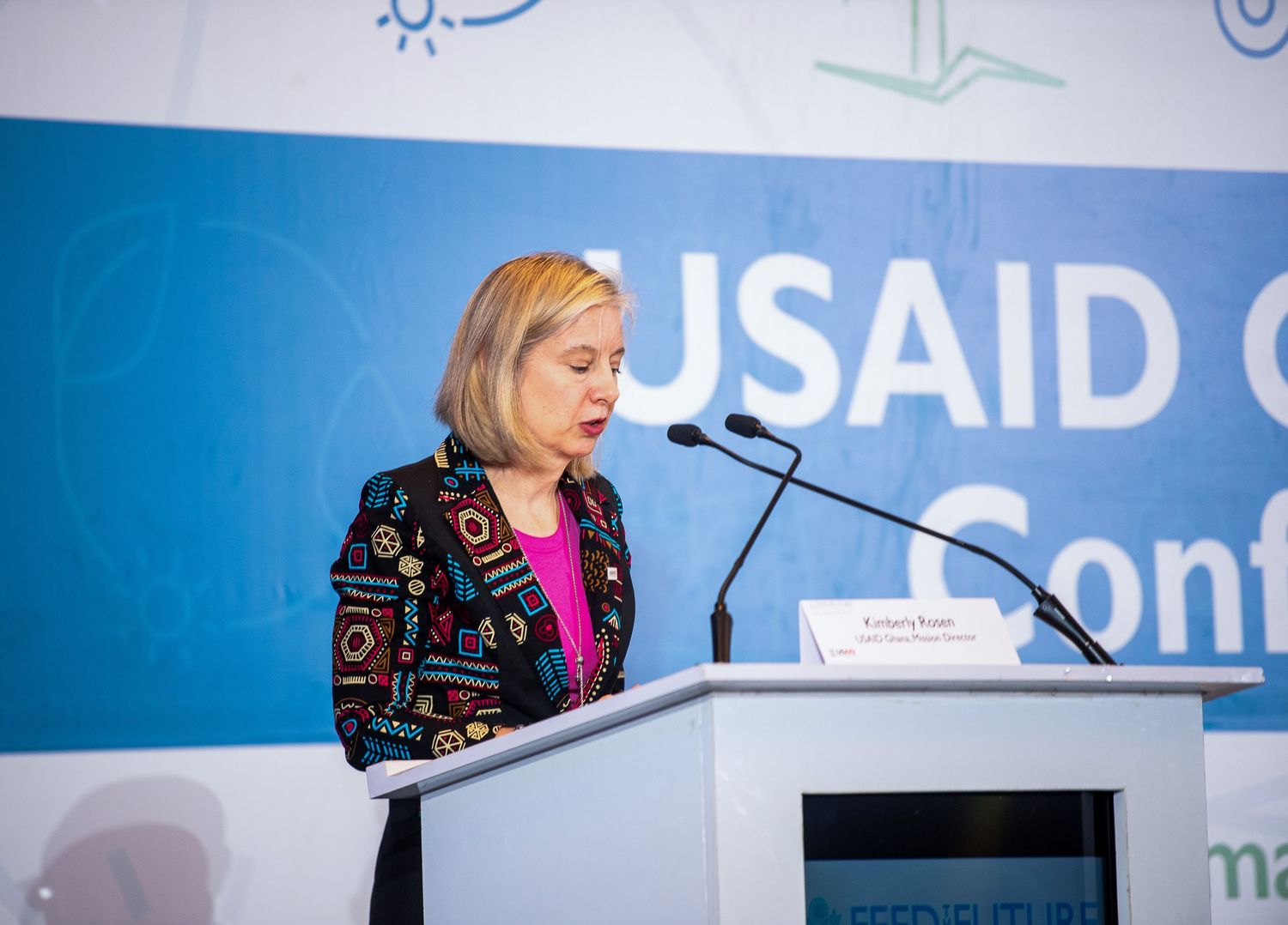 A speaker presents at a podium during a USAID conference with blue backdrop and organizational branding.