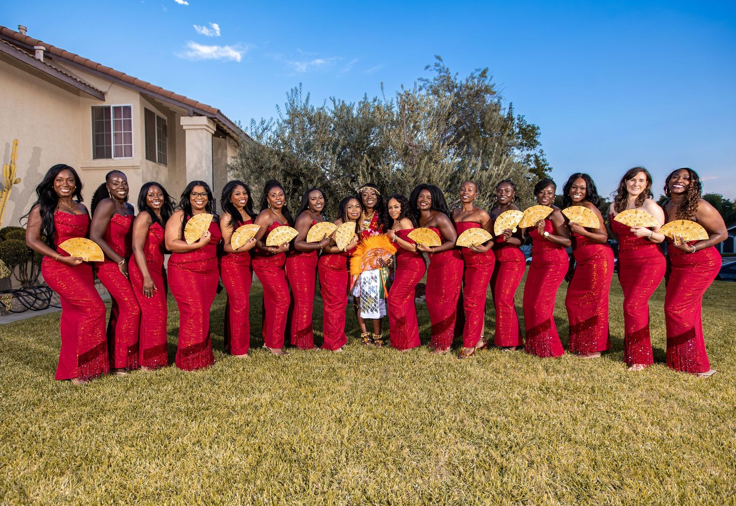 A group wearing matching red dresses poses together on a grassy hillside against a bright blue sky.