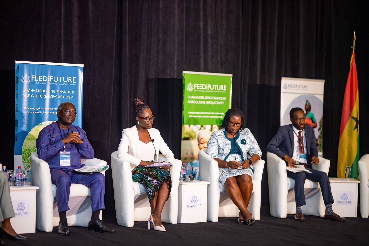 Panelists seated in white chairs on stage during a business conference with banners and flags visible in background.