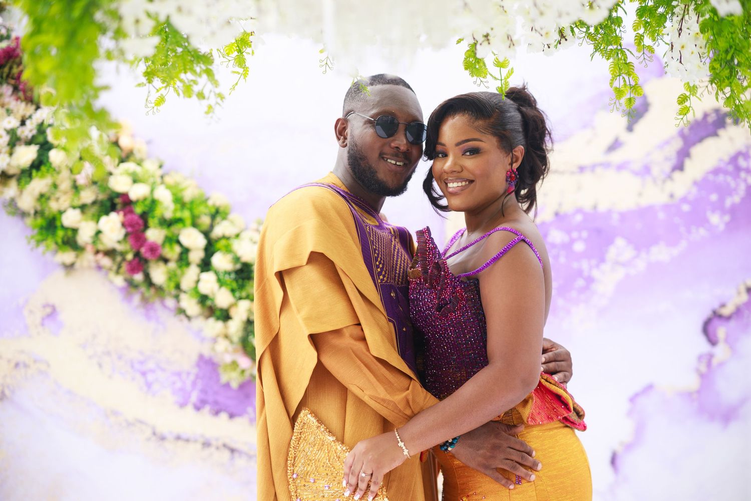 A couple in vibrant yellow and burgundy formal attire embrace while standing under flowering vines in an outdoor setting.