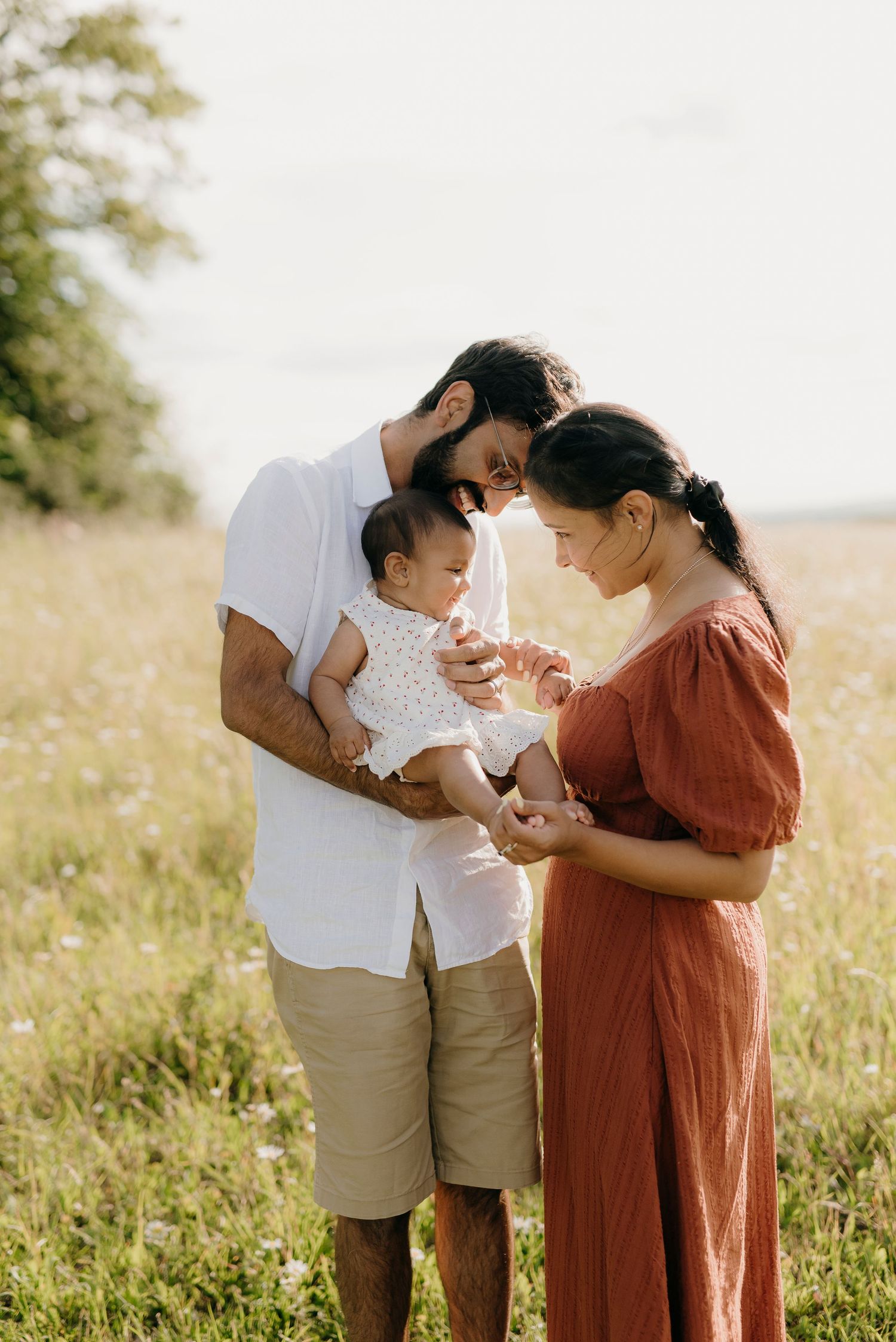 A young family stands in a sunny field, parents lovingly gazing at their newborn baby in a warm outdoor setting.