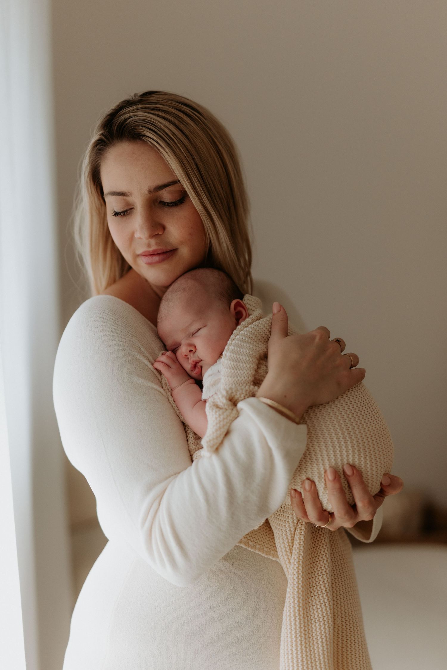 A young mother in a white dress tenderly holds her sleeping newborn baby close to her chest.