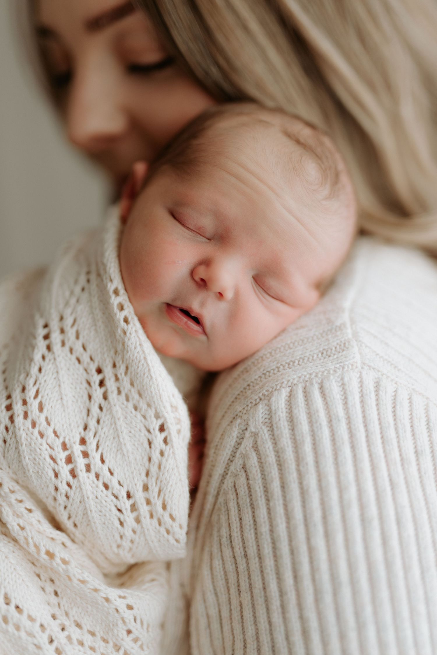A peaceful newborn baby sleeping wrapped in a cream knit blanket, cradled in a mother's arms.
