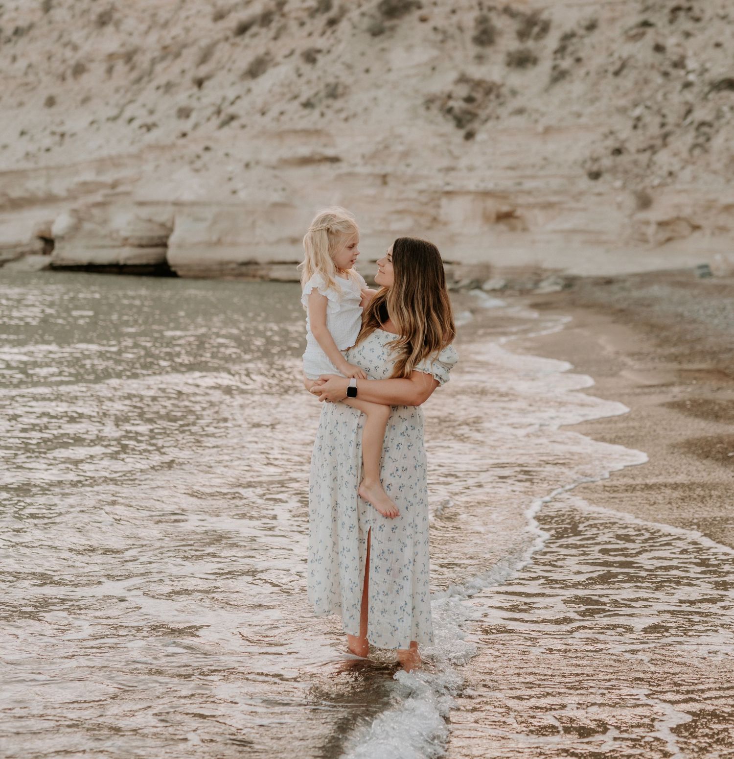 A mother holds her young daughter while standing in shallow ocean waves near a sandy cliff beach.