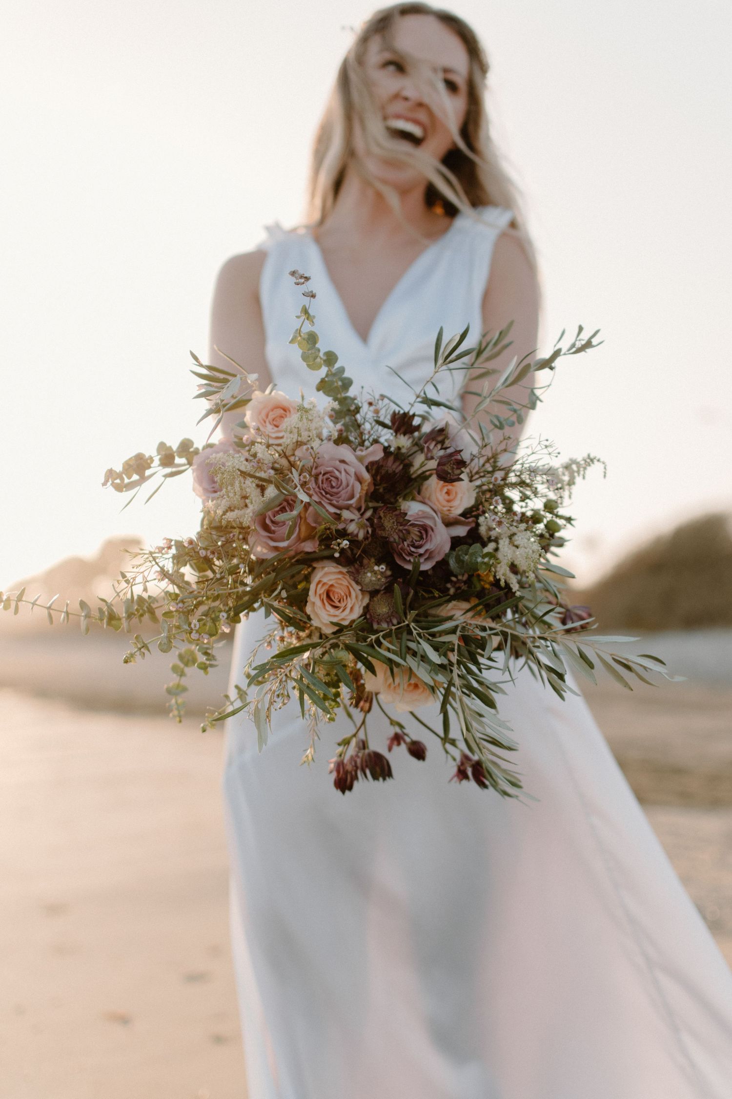 Laughing bride in white gown holds lush bouquet of pink roses and greenery on a sunlit beach.