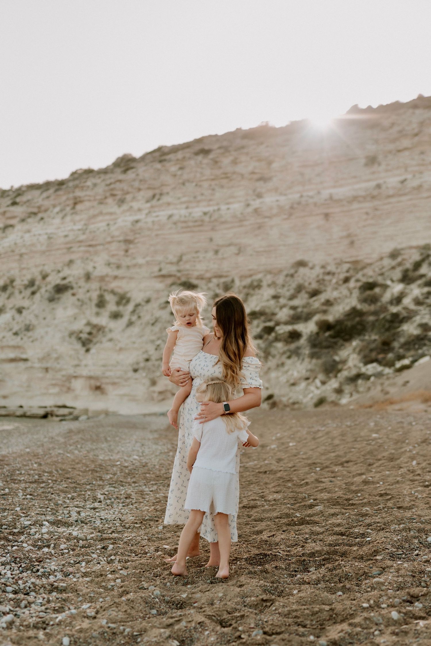 A mother in a white dress holds her toddler while another child hugs her on a rocky beach at sunset.