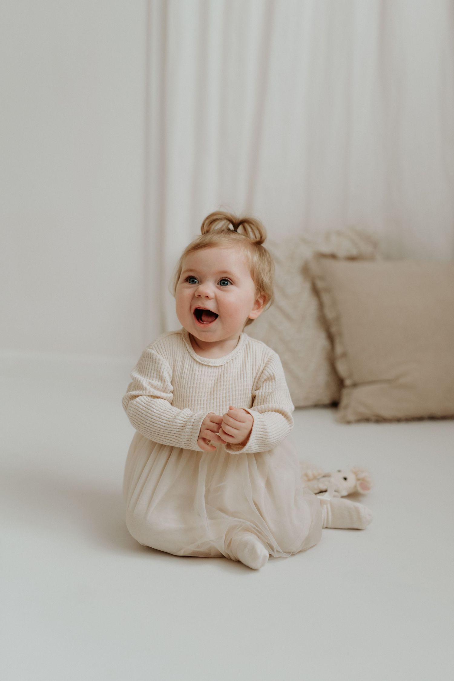 Happy baby girl in cream knit romper sitting on white floor, laughing with hands clasped together.