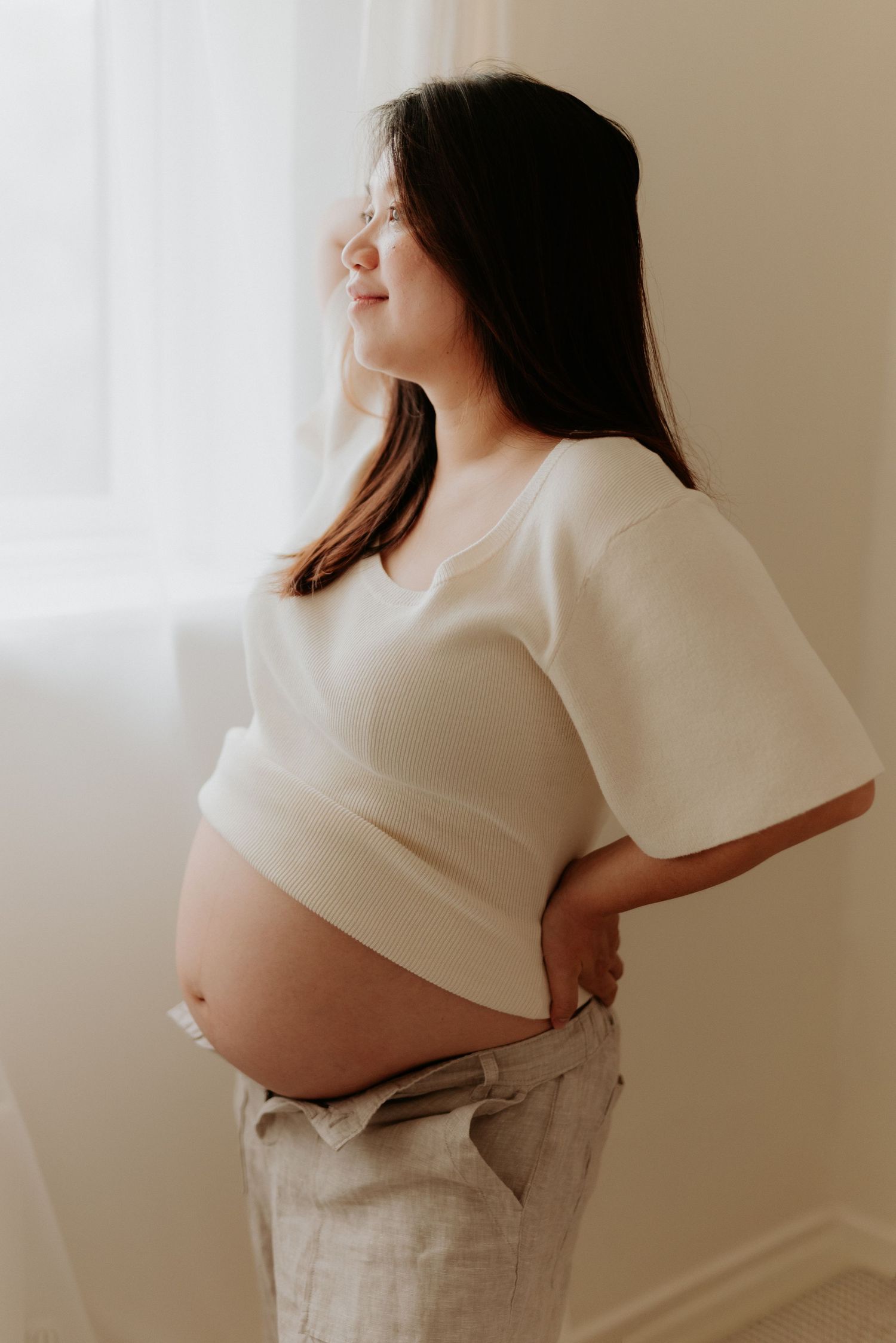 Pregnant woman in white top holding her baby bump while gazing out a window in soft natural light.