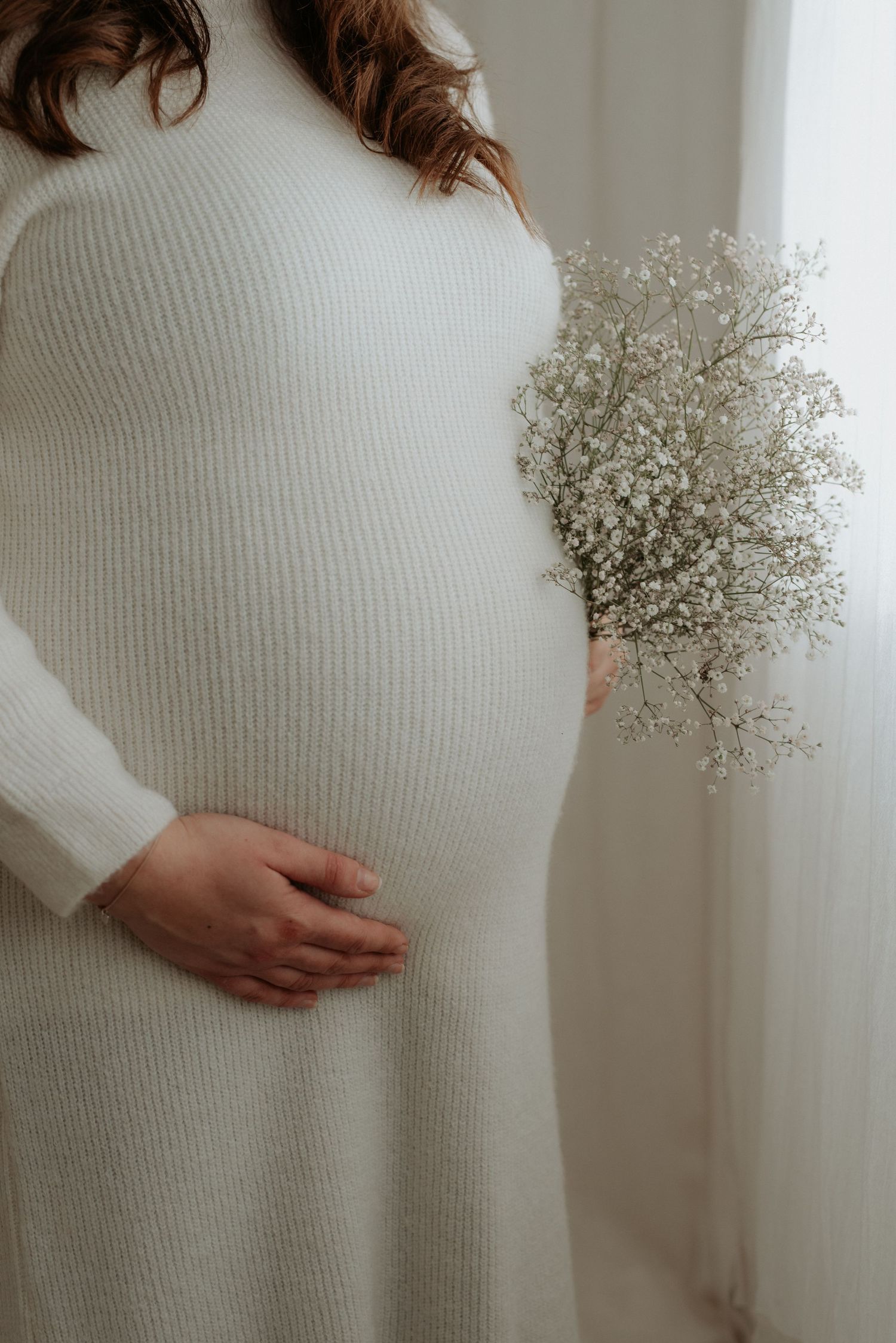 Pregnant woman in white ribbed dress holding baby's breath flowers, gently cradling her baby bump near a window.