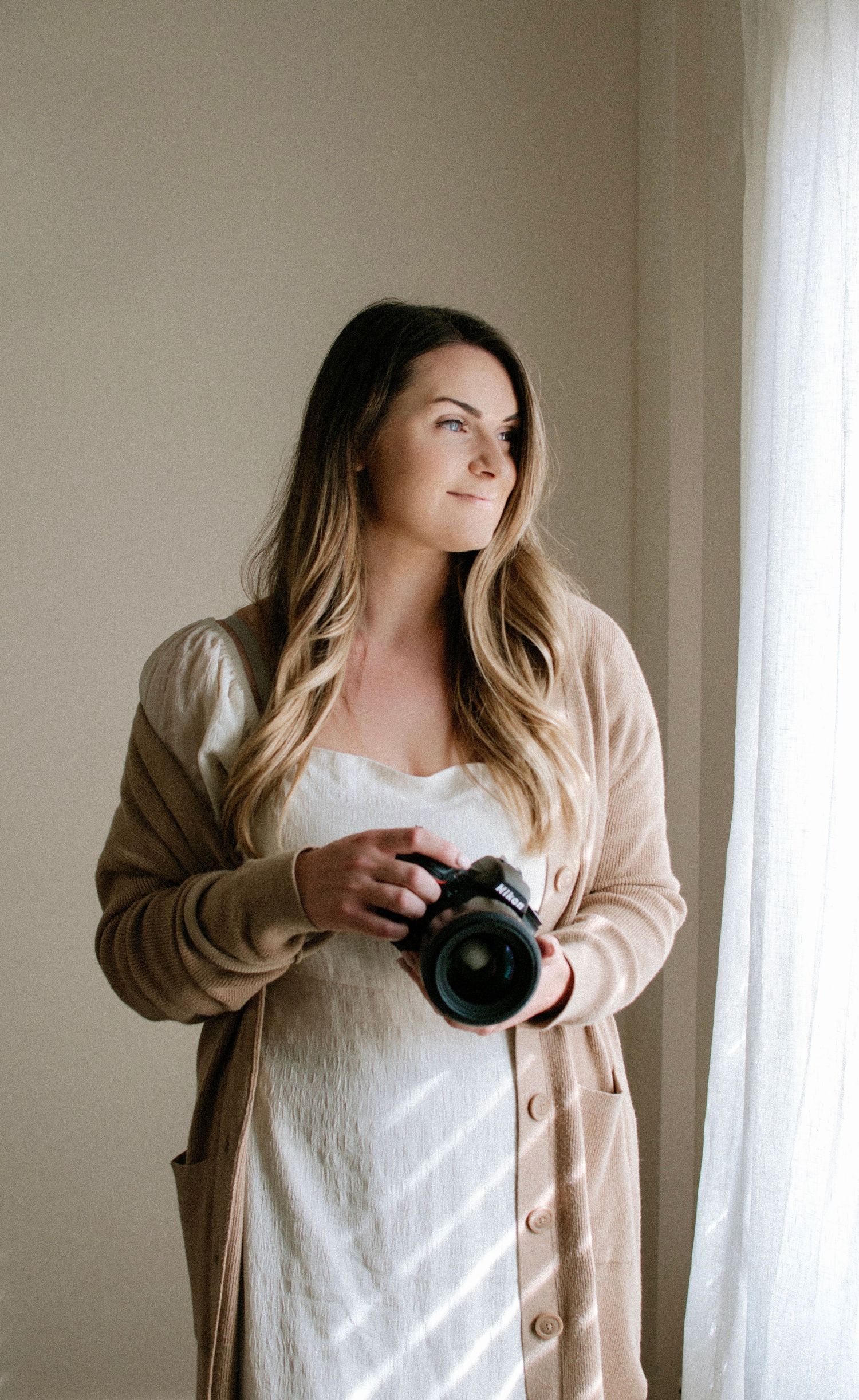 Woman in beige cardigan holding a DSLR camera, gazing thoughtfully toward natural window light.