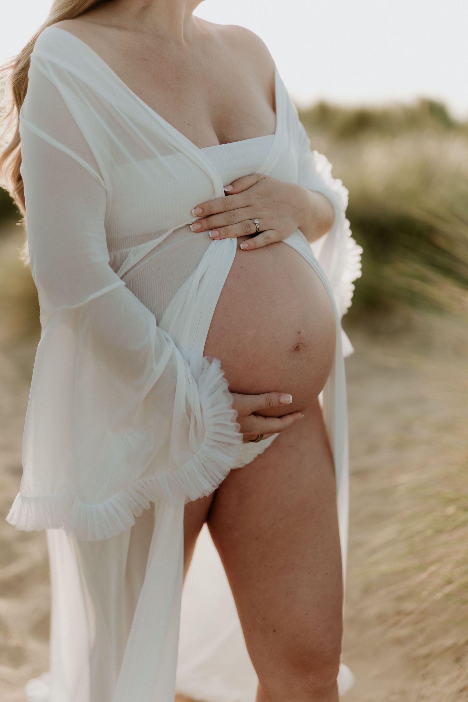 Pregnant woman in white sheer robe holding baby bump outdoors on sandy beach during golden hour.