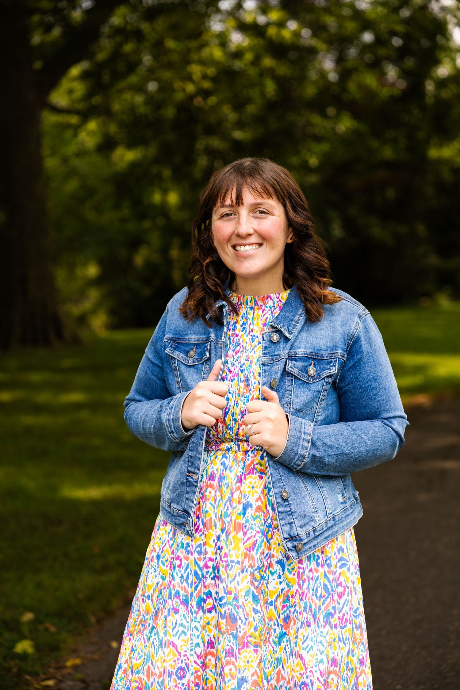 Smiling woman in floral dress and denim jacket standing in a lush green park pathway.