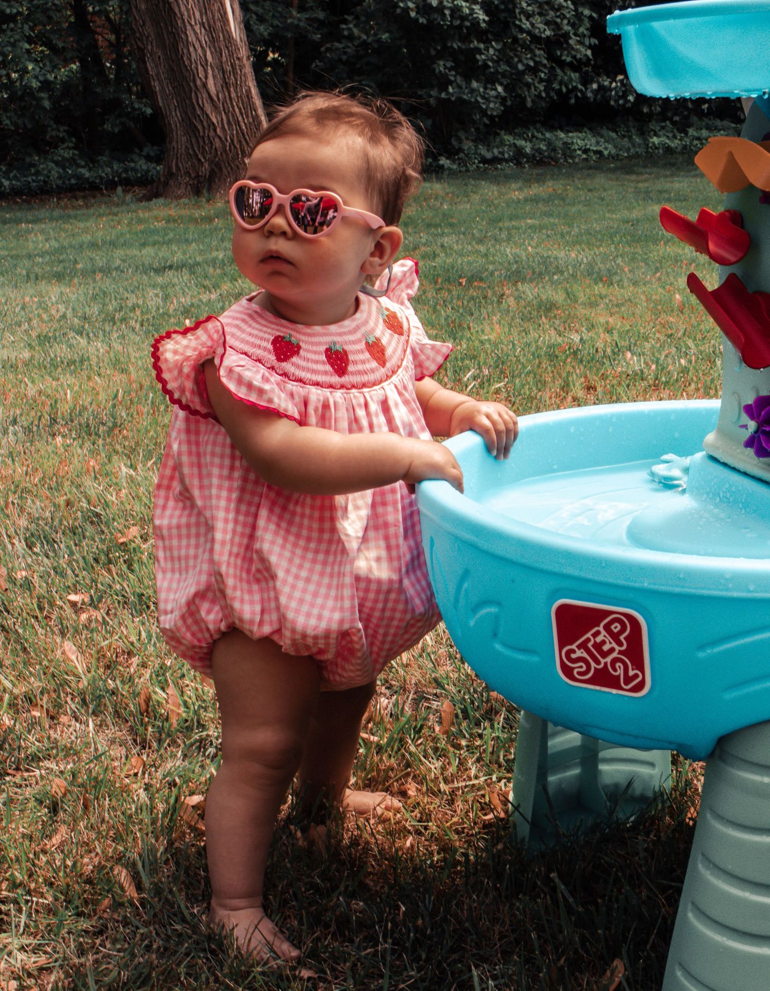A child plays with a bright blue water table outdoors while wearing a pink gingham outfit.