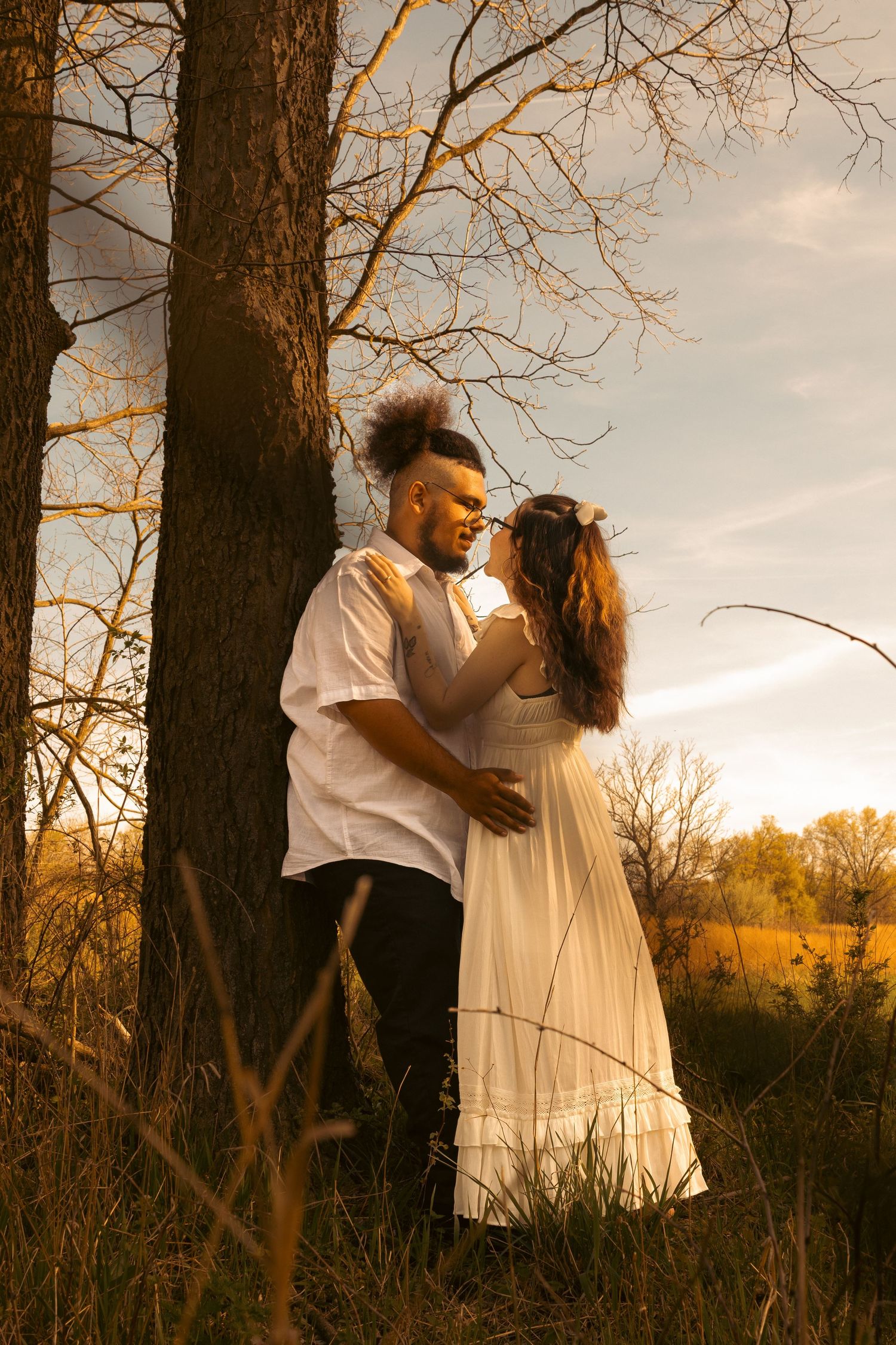 Romantic couple embracing under a tree at golden sunset, woman in white dress, outdoor nature photography.