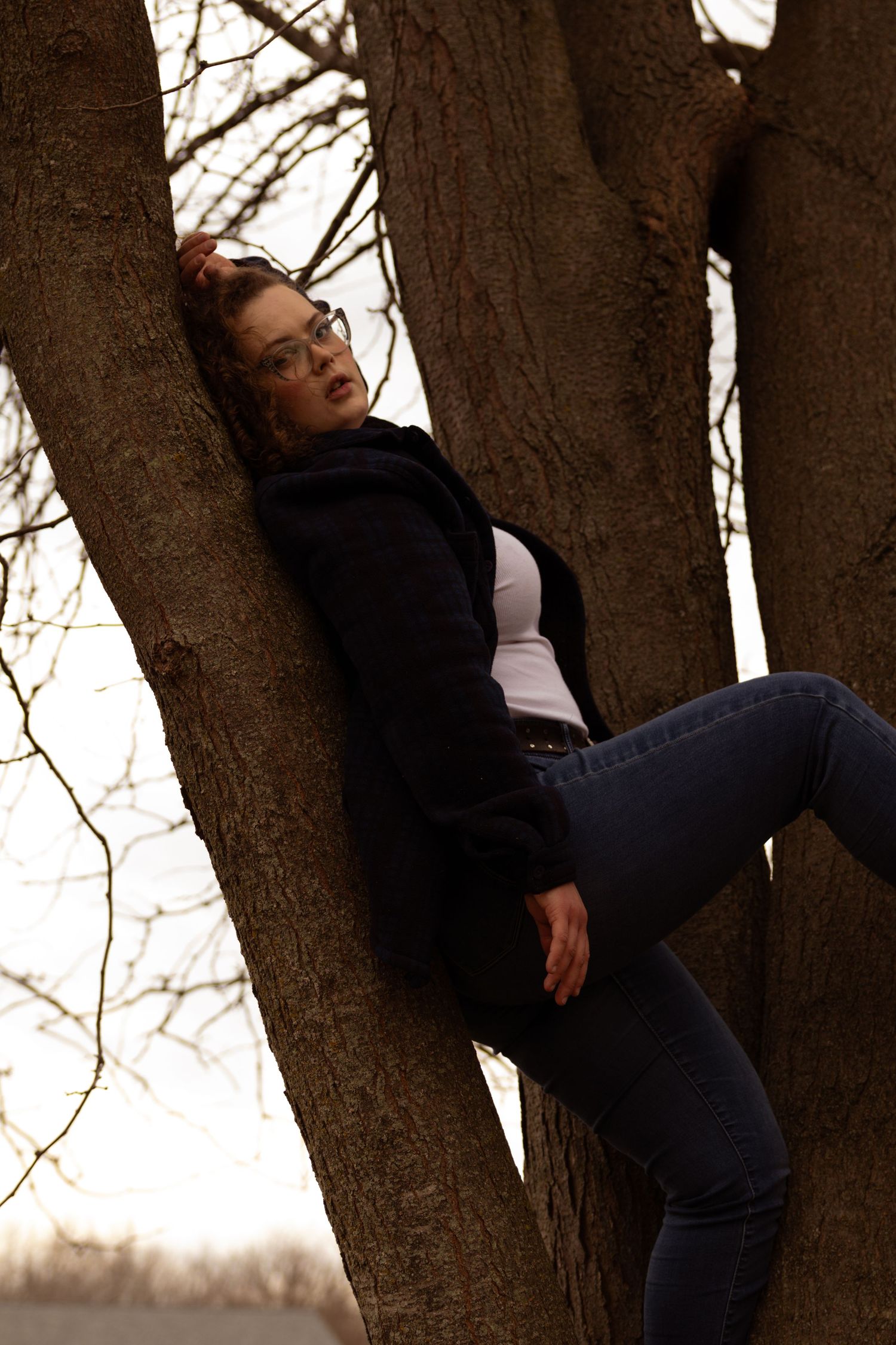 Young woman in jeans and black jacket leaning against tree trunk outdoors, wearing glasses with hair up.