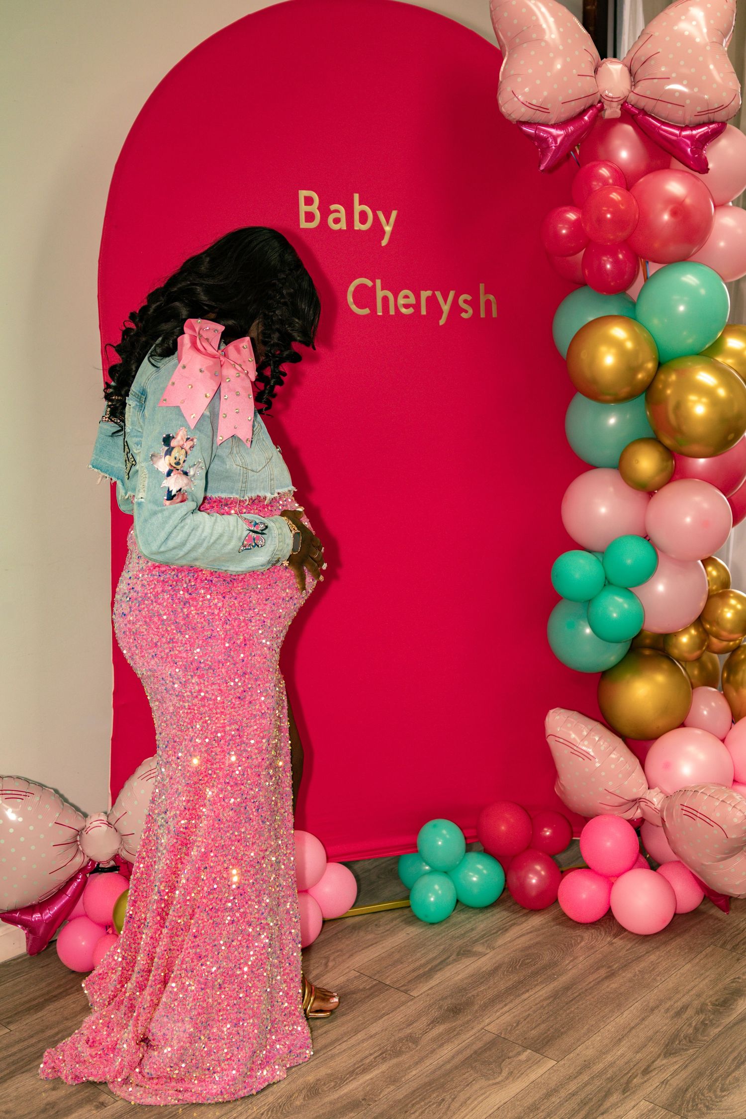 Pregnant woman in pink sequin gown at Baby Cherysh shower with colorful balloon arch backdrop.